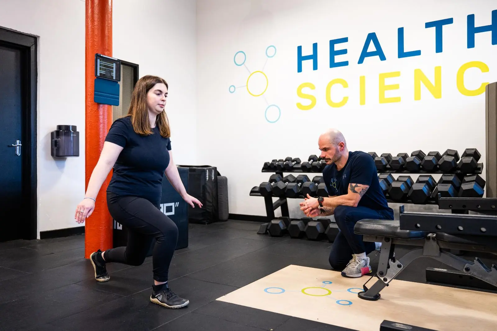 Female athlete performing a reverse lunge with a personal trainer in a gym setting.