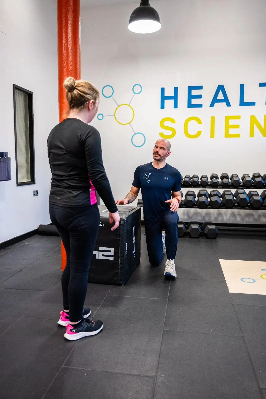 KA3_2997 A female client standing and talking with a male trainer in front of the Health By Science logo wall, illustrating a personalized consultation or assessment.