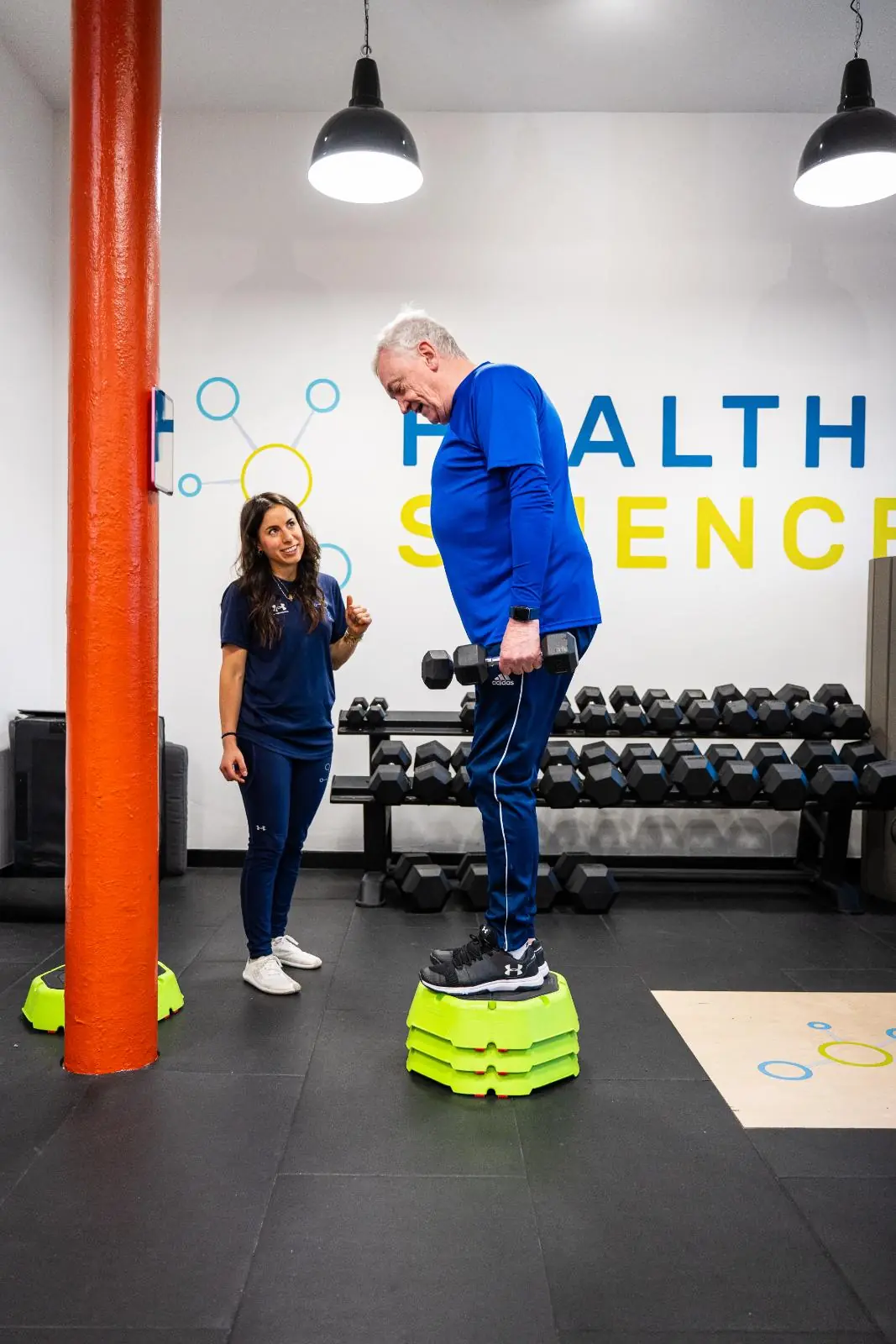 KA3_3060 A female Health By Science physiotherapist talking with an older male client who is holding a dumbbell, demonstrating a supportive and inclusive training environment.