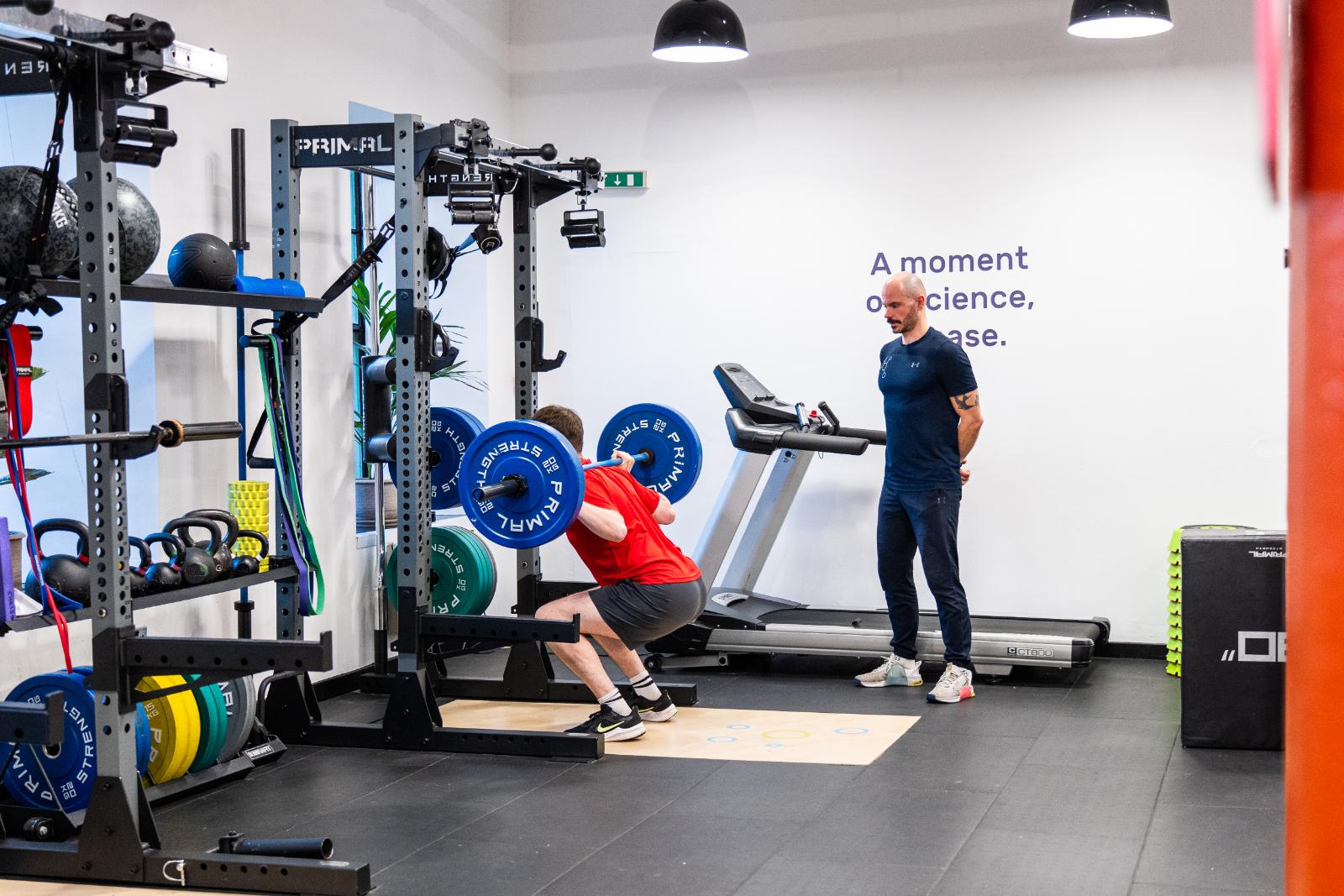 A man performing a back squat with a barbell while a personal trainer edinburgh observes his form at Health By Science Leith.