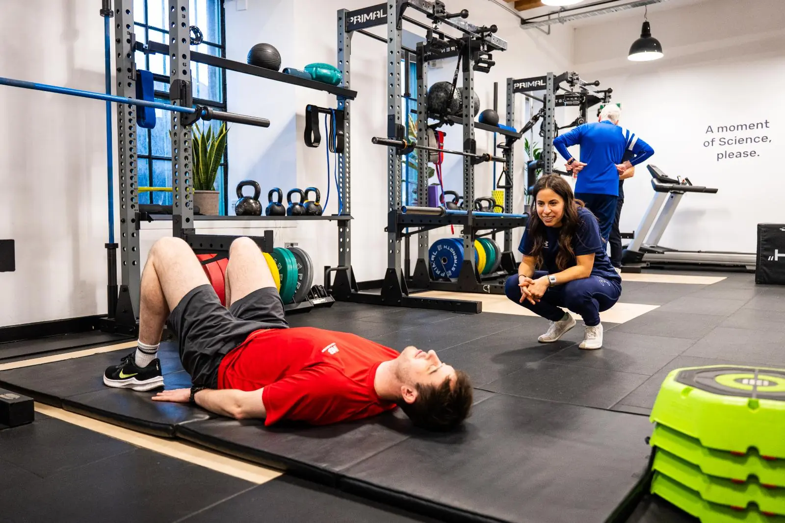 KA3_3326 A male client lying on a gym mat performing a bridge exercise while a female trainer crouches nearby to assess his movement and form.