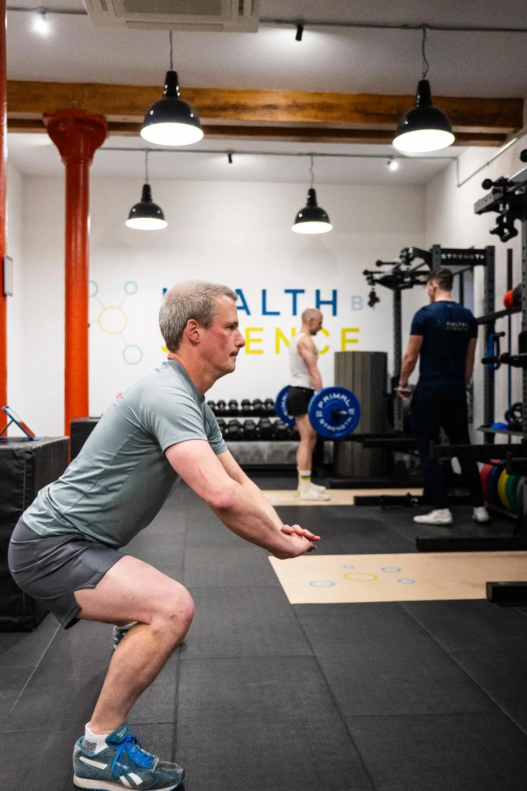 KA3_3451 Health by Science coach monitoring a patient performing a weighted squat to build lower body strength and joint resilience.