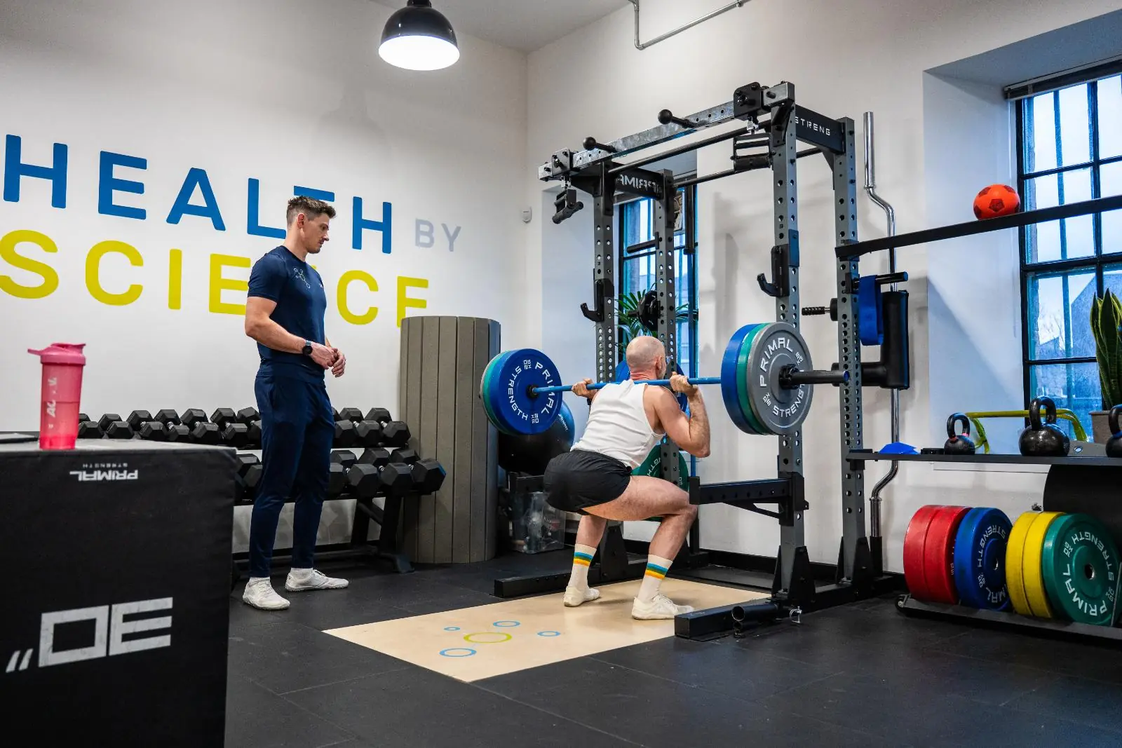 A man performing a heavy barbell back squat in a high-performance gym while a coach monitors his form.