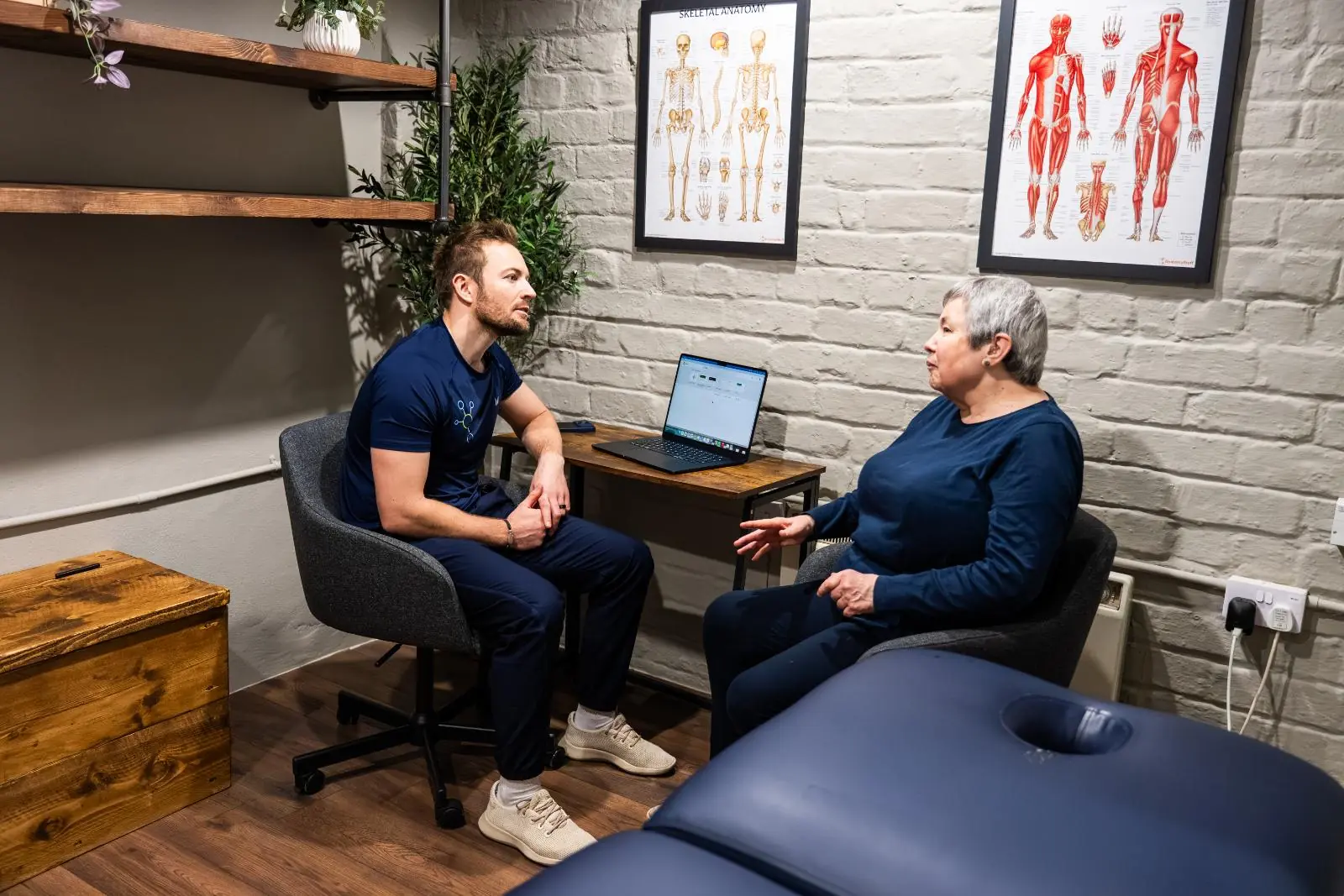 A male physiotherapist in blue scrubs conducting an initial consultation with a patient in a private clinic room.