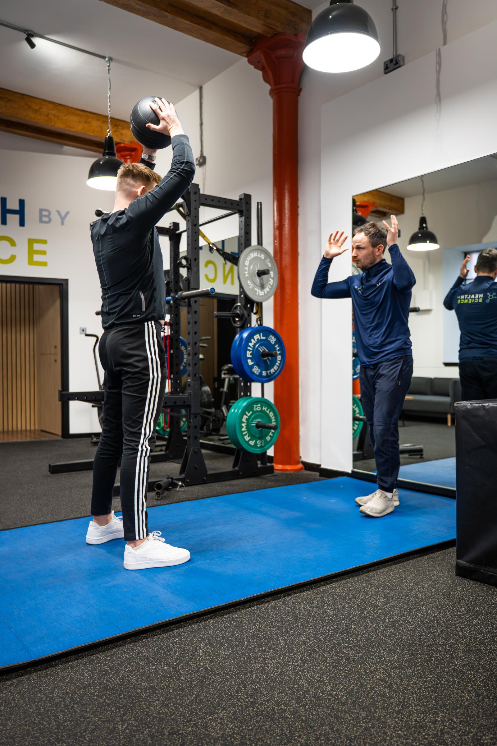 KA3_5139 Specialist providing technical feedback on overhead medicine ball positioning to ensure optimal shoulder mechanics and core engagement.