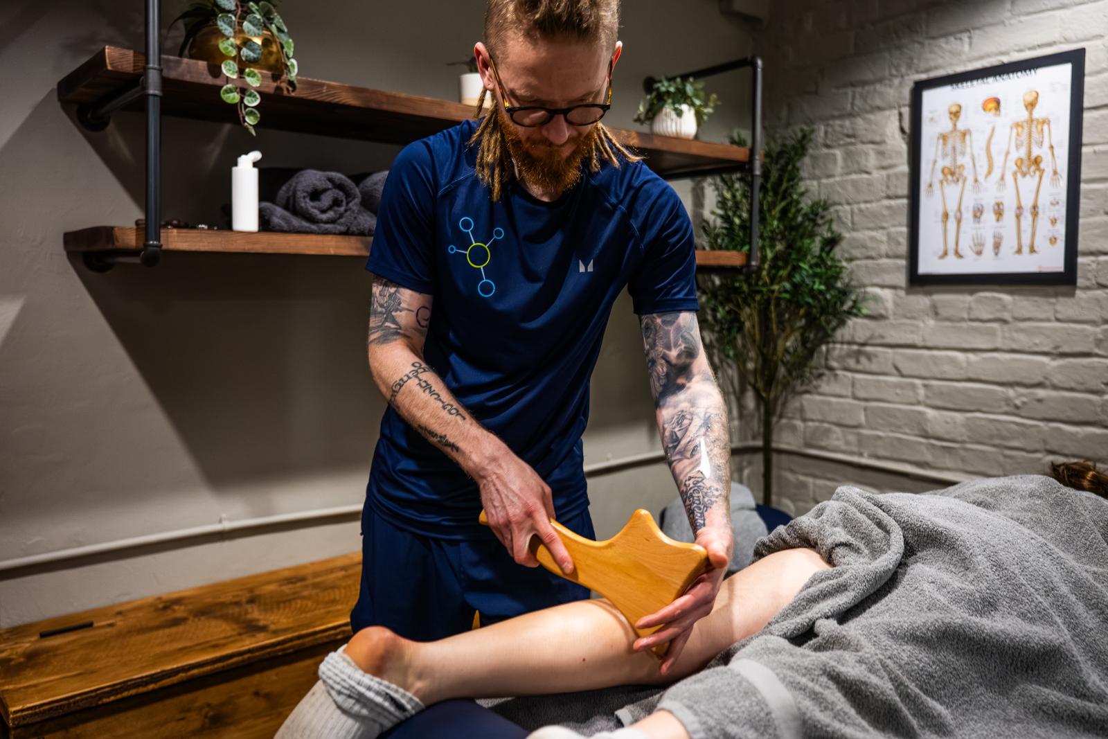 KA3_5758 A male sports massage edinburgh therapist using a specialised wooden massage tool for deep tissue muscle recovery on a patient's leg.